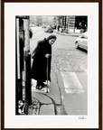 An elderly woman in a headscarf and long coat uses a cane to step off a curb onto a cobblestone street at a crosswalk. Cars and pedestrians are visible in the background. This black and white photo is presented as framed wall art for conscious interior design.