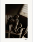 A man sits on a wooden chair indoors, resting his arm on his knees and looking to the side. This black and white image with soft lighting and a blurred background is ideal as framed wall art for mindful home decor. An artist’s signature marks the lower right corner.
