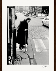 An older woman with a cane crosses a cobblestone street at a crosswalk in this black and white photo. Wearing a dark coat and headscarf, she moves past cars and pedestrians—perfect as framed wall art for conscious interior design.