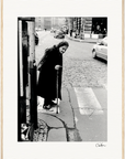 An elderly woman in a headscarf and long coat uses a cane to cross a cobblestone city street near a telephone booth. The black and white image makes striking framed wall art for conscious interior design. Cars are visible in the background.