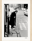 An elderly woman in dark clothing uses a cane to cross a cobblestone street at a crosswalk. Cars and buildings are visible in the background of this black-and-white inspirational wall art, perfect for any space as framed wall art.
