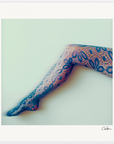 A single outstretched human leg is shown against a pale background, wearing black lace-patterned tights with a floral design. The image has a soft focus and would make a striking fine art print for conscious interior design. A signature is visible in the bottom right corner.