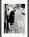 An elderly woman with a cane crosses a cobblestone street at a crosswalk, dressed in a long dark coat and headscarf. Cars and buildings are visible in the black-and-white image, ideal as a fine art print for mindful home decor.