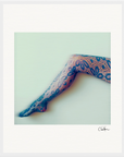 A person’s leg is extended against a pale background, wearing blue patterned lace tights with large floral designs. This soft-focus image is perfect as inspirational wall art or mindful home decor, complete with a signature in the bottom right corner.