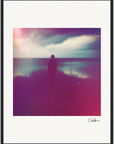 A silhouetted figure stands near the edge of a calm body of water under a cloudy sky, with purple and pink light effects and faint film grain texture. This fine art print is framed in a gallery-quality frame and signed in the lower right corner.