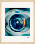 A close-up, slightly distorted view of a washing machine door with the Electrolux brand label visible, surrounded by blue and teal reflections, framed with light wood—an inspirational wall art piece for conscious interior design.