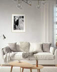 A bright living room with a white sofa, neutral-toned pillows, a floor lamp, and a small wooden table. A fine art print in a light frame hangs above the sofa, adding mindful home decor to the space. White curtains cover the window on the right.