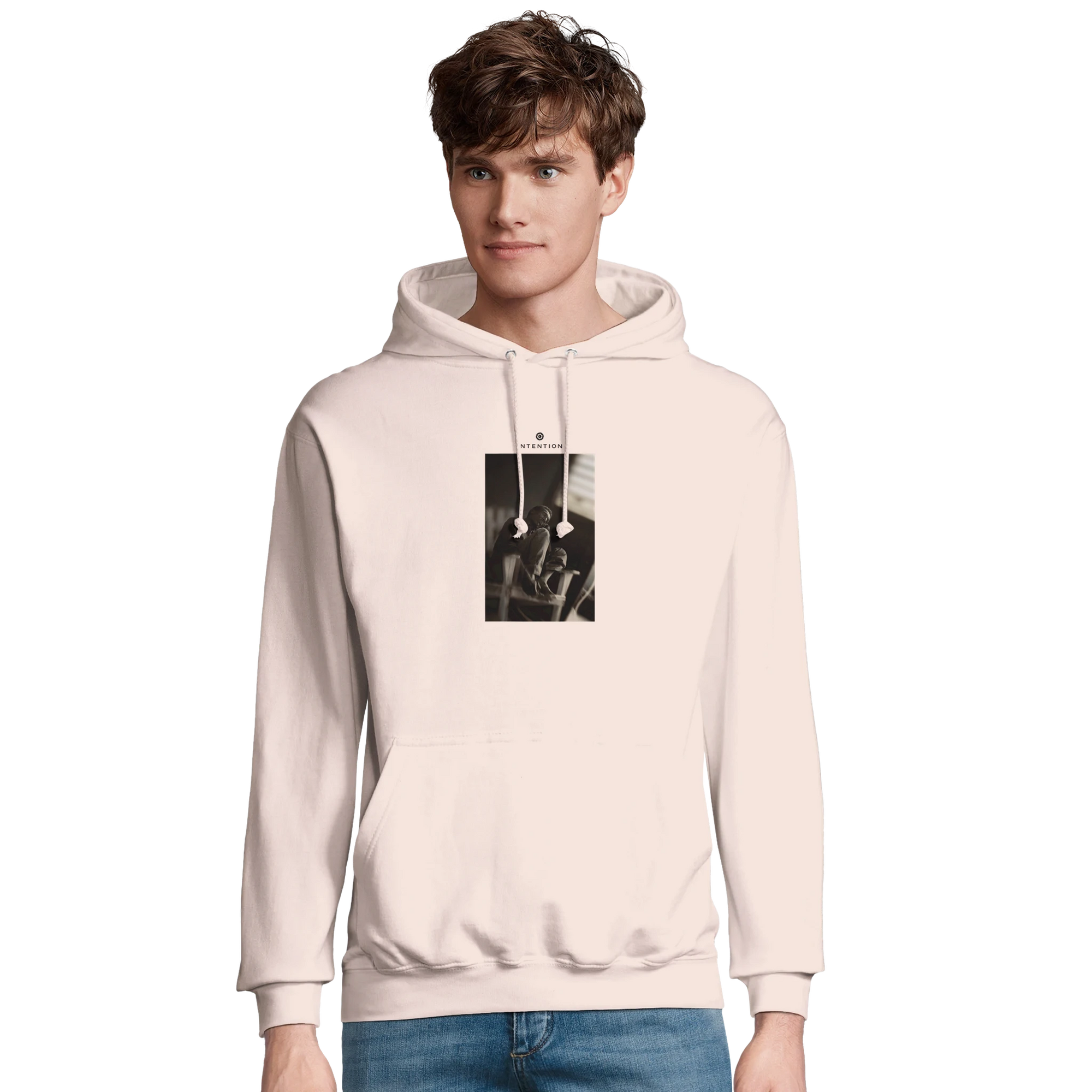 A young man with short brown hair wears a light-colored hoodie featuring a black-and-white art print on the chest. He is standing against a plain, light background and wearing blue jeans.