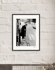 A framed black-and-white fine art print hangs on a white brick wall. The photo shows an elderly person with a cane standing on a cobblestone street corner, inspiring mindful home decor with its timeless city scene.