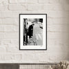 A framed black-and-white fine art print hangs on a white brick wall. The photo shows an elderly person with a cane standing on a cobblestone street corner, inspiring mindful home decor with its timeless city scene.
