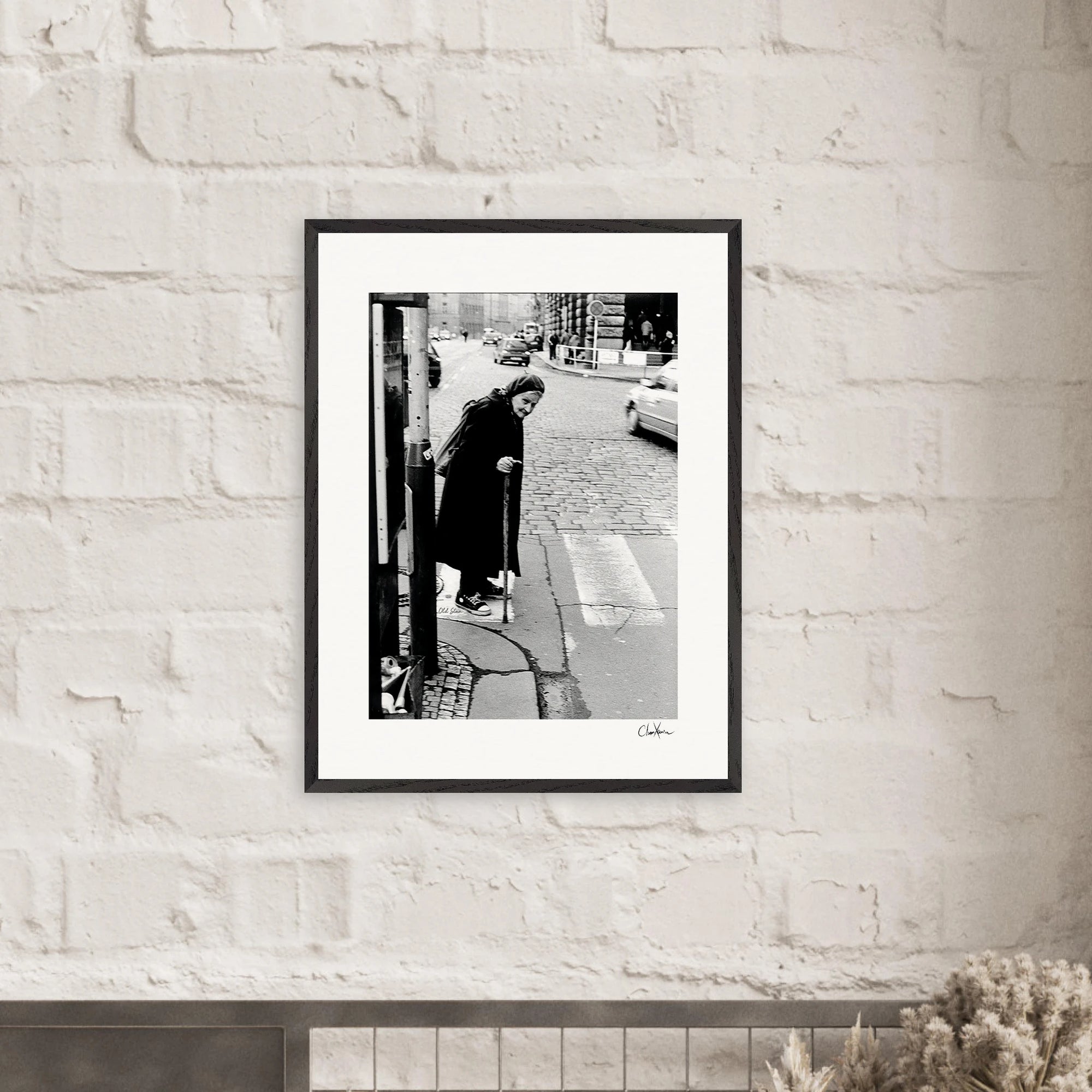 A framed black-and-white fine art print hangs on a white brick wall. The photo shows an elderly person with a cane standing on a cobblestone street corner, inspiring mindful home decor with its timeless city scene.