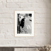 A gallery-quality frame showcases this black-and-white photograph, depicting an elderly person with a cane at a city street corner. The striking image adds inspirational wall art to any conscious interior design.