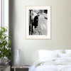 A framed wall art black-and-white photograph of an elderly person with a cane stepping onto a street hangs above a white bed with pillows; a plant and white vase sit on a small table nearby, creating mindful home decor.