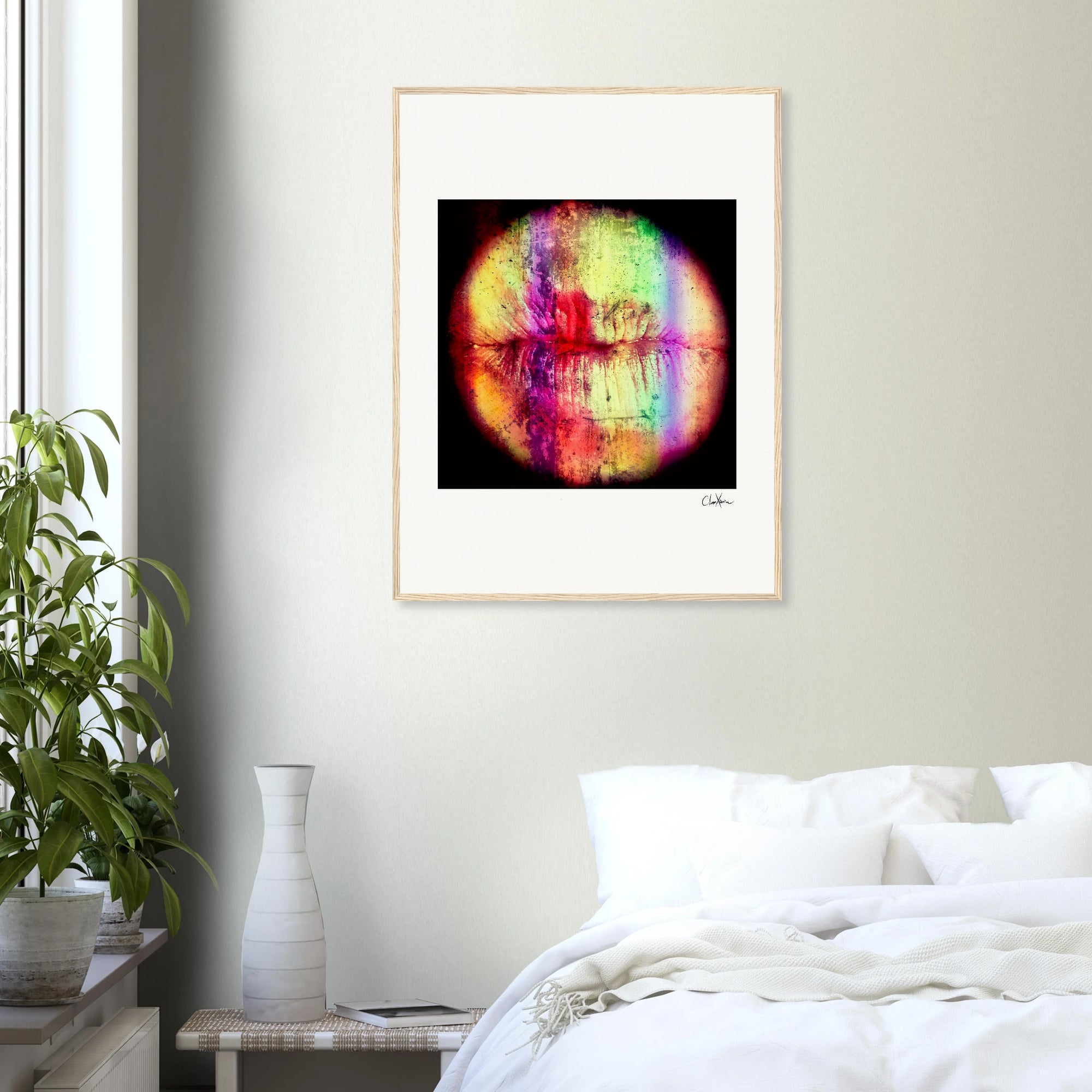 A framed fine art print with vibrant, blended colors hangs on a light-colored wall above a bed with white bedding. A green potted plant and a white vase rest on a small table beside the bed, creating an inspirational wall art display.