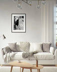 A modern living room with a white sofa, neutral pillows, a throw blanket, a round wooden coffee table, and wall art in a gallery-quality frame above the sofa. White curtains and large windows bring in natural light.