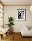 A minimalist bedroom with a low bed, wooden desk and chair, a potted plant, and a paper lantern light. A fine art print in a black frame hangs on the wall above the bed, adding mindful home decor to the serene space.
