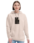 A person with short brown hair wears a light beige hoodie featuring an art print graphic on the chest, paired with light blue jeans. The look reflects conscious fashion against a plain white background.