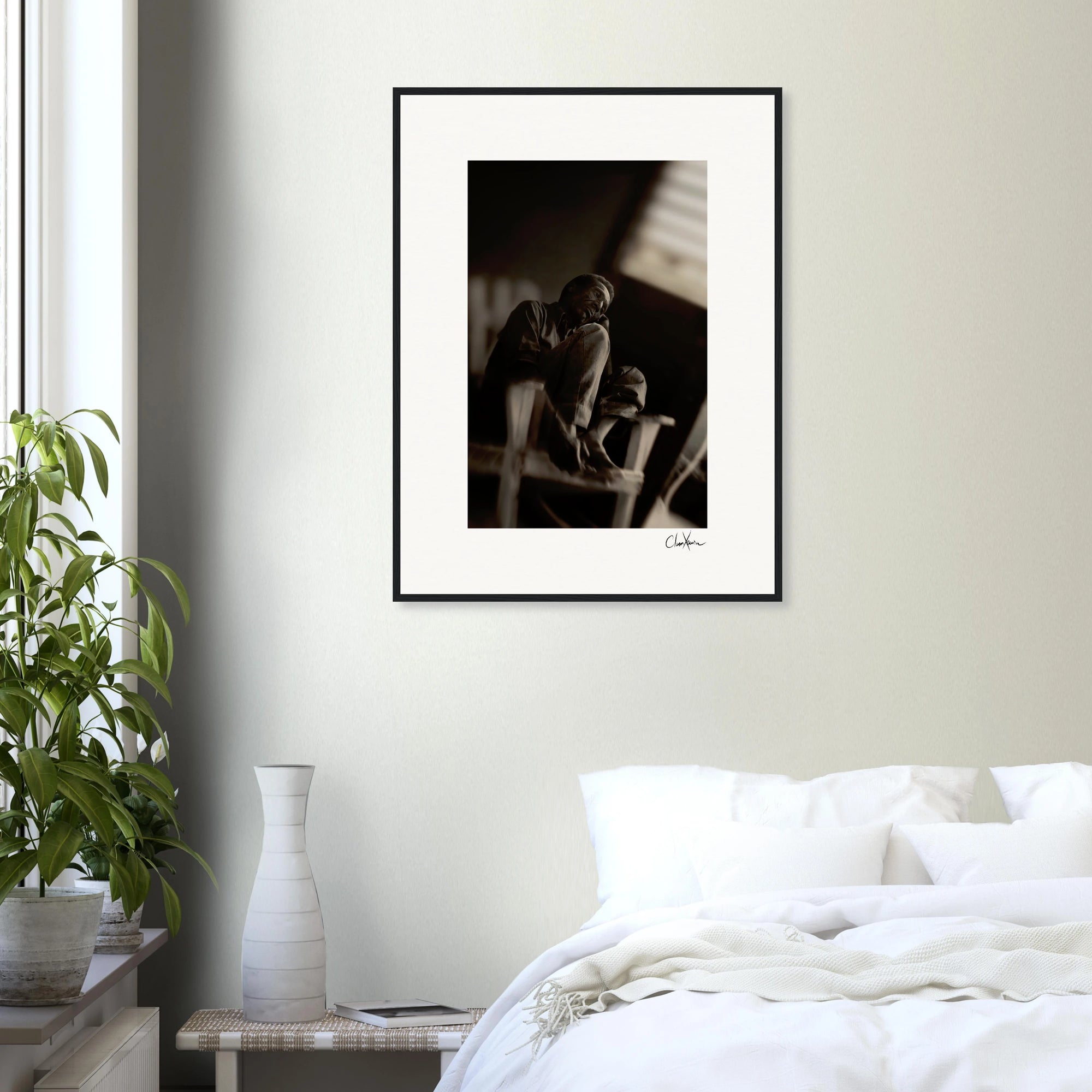 A framed wall art black-and-white photograph hangs on a light-colored wall above a white bed. A green potted plant and a white vase rest on the bedside table, adding mindful home decor touches.