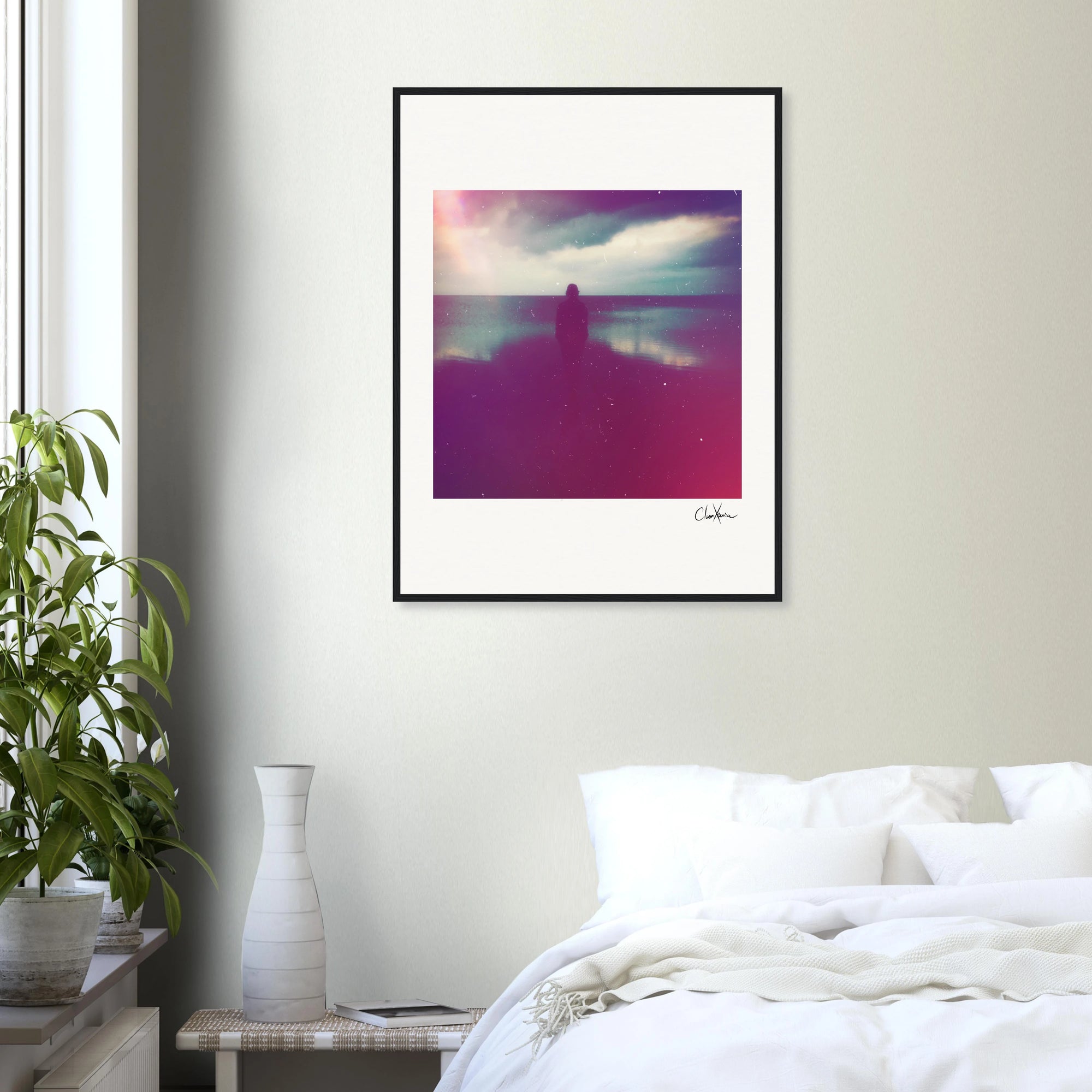 A framed fine art print hangs on a beige wall above a white bed. The image features a silhouette of a person and a colorful, blurry landscape. A potted plant and white vase sit on the bedside table, perfect for conscious interior design.