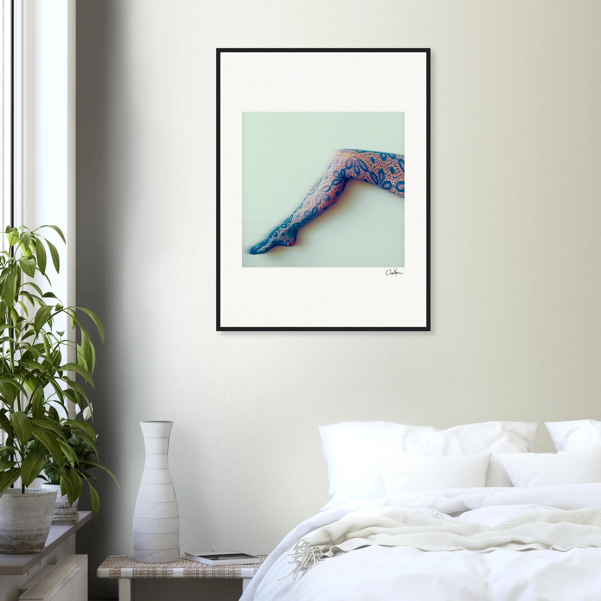 A minimalist bedroom features a white bed, a leafy green plant in a white pot, and inspirational wall art—an elegant fine art print of a tattooed leg framed against a pale background.