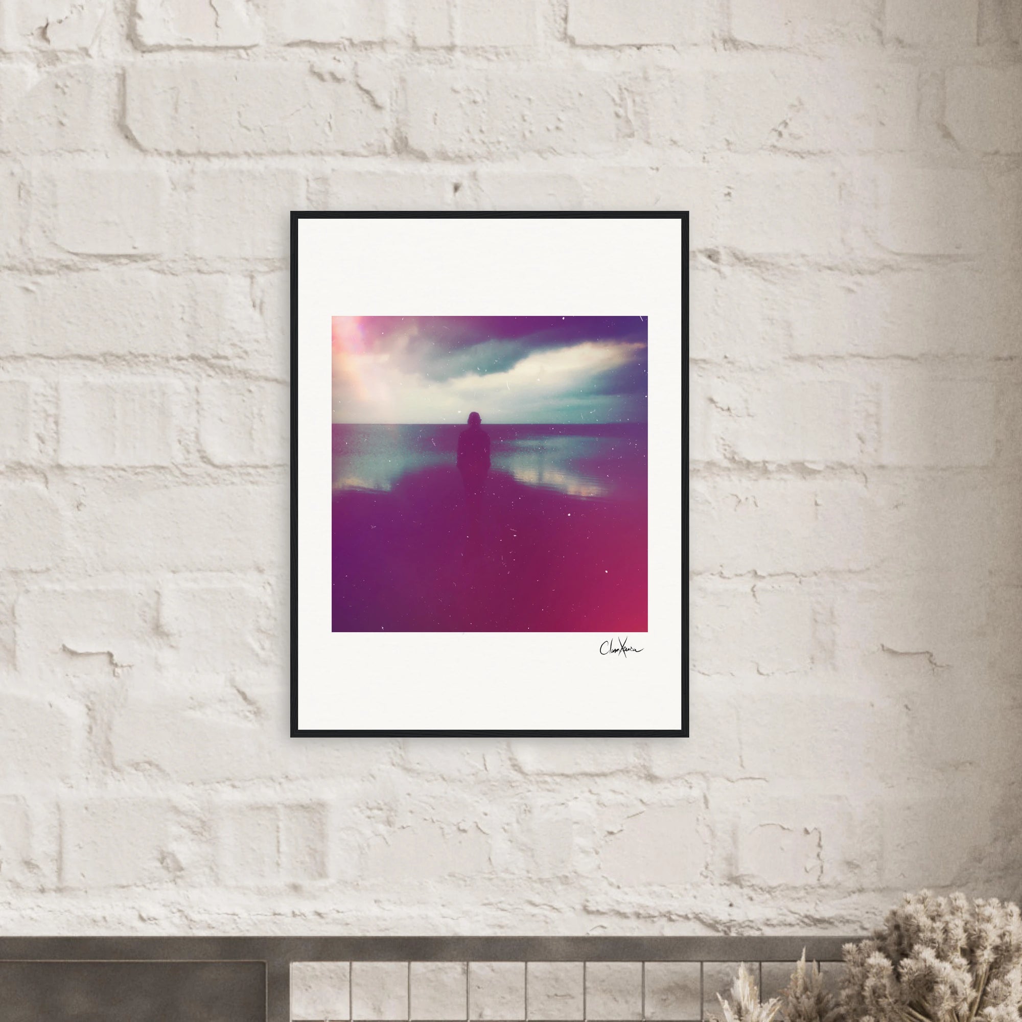 A framed photograph hangs on a white brick wall, serving as inspirational wall art. The gallery-quality frame surrounds a dreamy image of a silhouetted figure by water with purple and pink hues, perfect for conscious interior design.