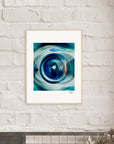 A framed fine art print featuring a close-up image of a washing machine door hangs on a white brick wall above a dark shelf with dried flowers in the foreground, adding an inspirational touch to conscious interior design.