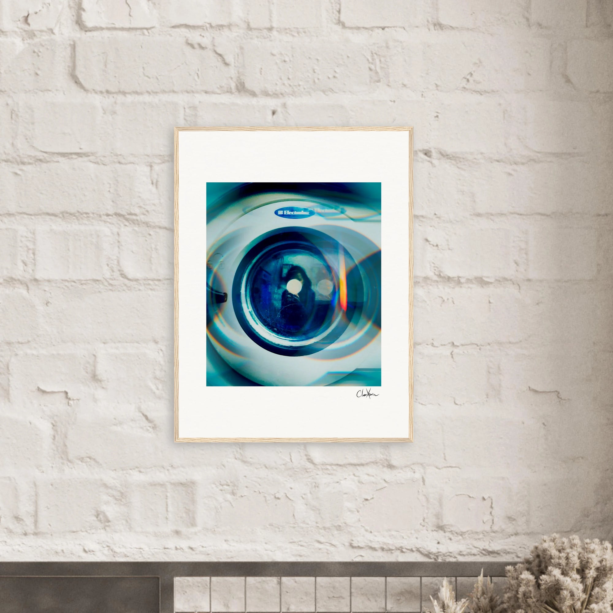 A framed fine art print featuring a close-up image of a washing machine door hangs on a white brick wall above a dark shelf with dried flowers in the foreground, adding an inspirational touch to conscious interior design.