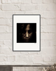A gallery-quality framed wall art photograph of a person's face with dark hair partially covering it hangs on a white brick wall above a shelf. The image has a dark, moody tone and is signed in the lower right corner.