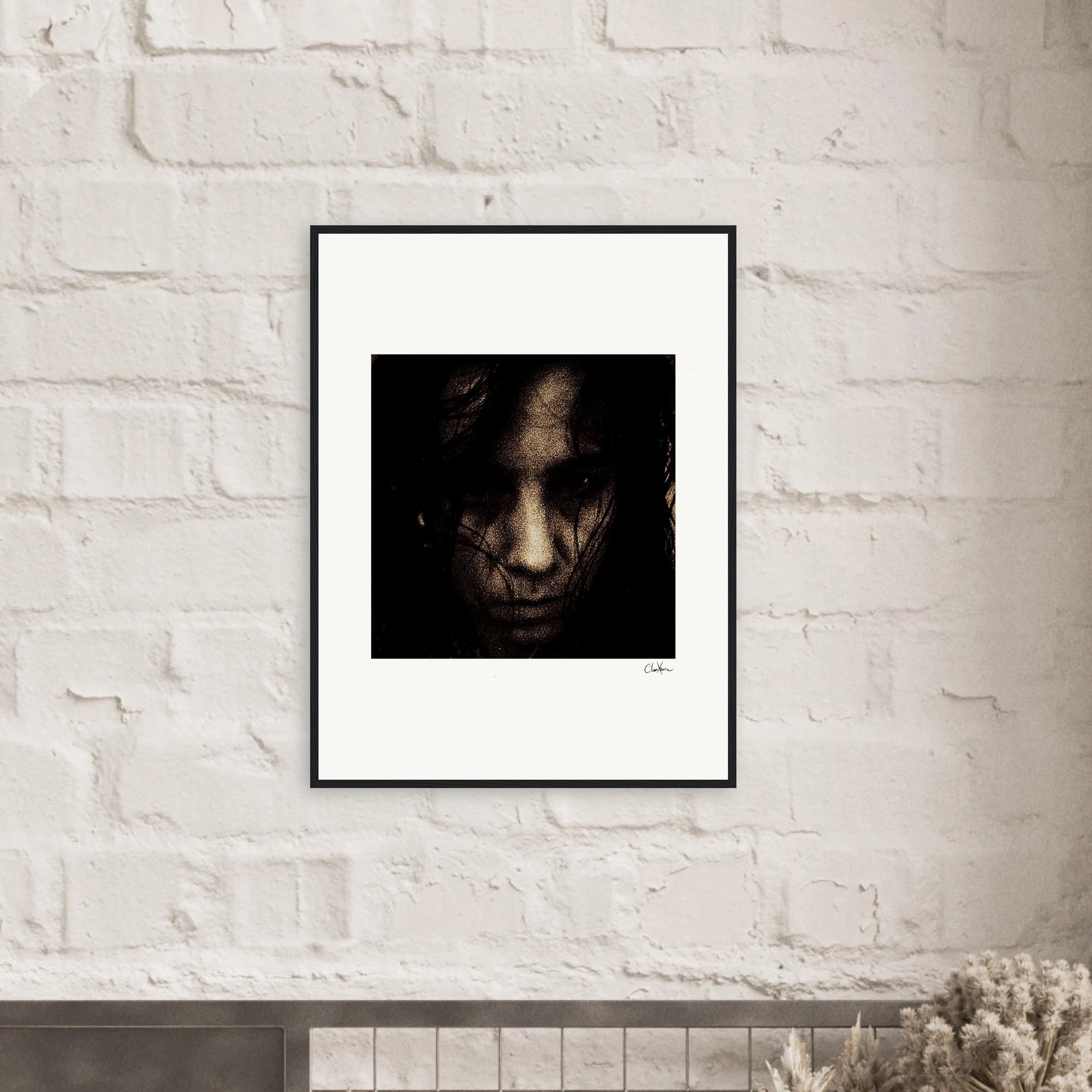 A gallery-quality framed wall art photograph of a person's face with dark hair partially covering it hangs on a white brick wall above a shelf. The image has a dark, moody tone and is signed in the lower right corner.