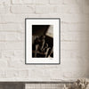 A fine art print in black and white, featuring a person sitting with knees drawn up, is displayed as framed wall art above a light-colored mantle adorned with a small plant and decorative objects—perfect for conscious interior design.