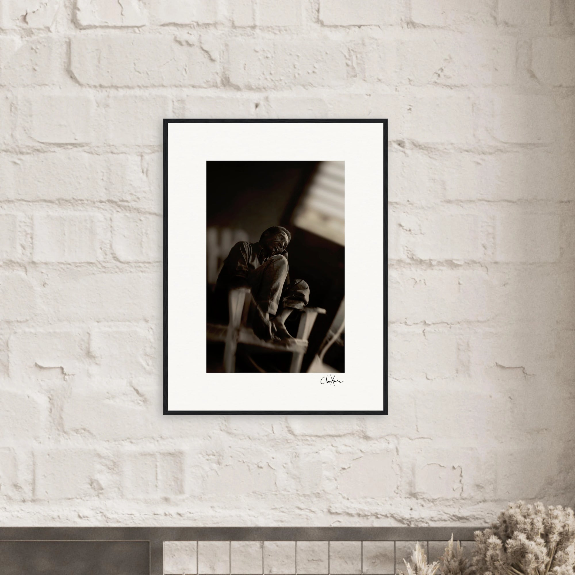 A fine art print in black and white, featuring a person sitting with knees drawn up, is displayed as framed wall art above a light-colored mantle adorned with a small plant and decorative objects—perfect for conscious interior design.
