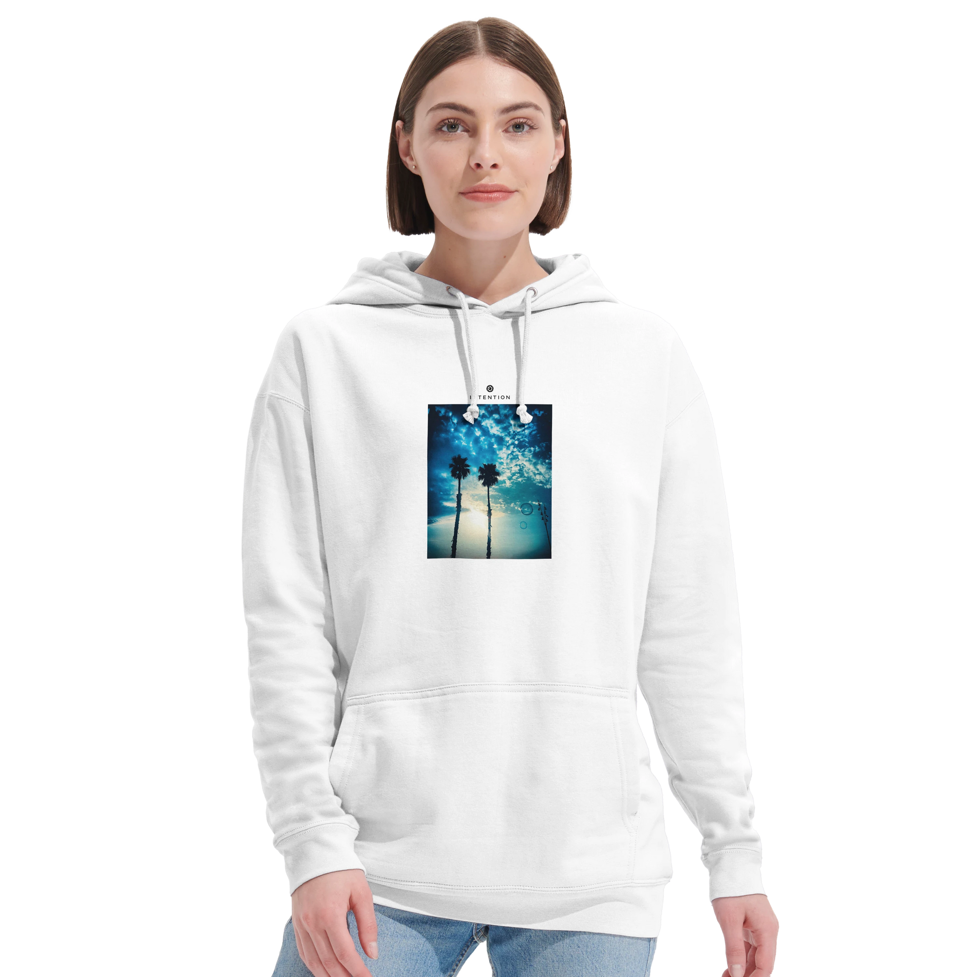 A person wearing a white hoodie from mindful apparel, featuring a printed image of palm trees and a blue sky with clouds on the front, stands against a plain white background.