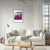 A modern living room with a white sofa, gray and beige cushions, a soft throw, floor lamp, and round wooden coffee table. Framed wall art in a gallery-quality frame hangs above the sofa, complemented by a large window with white curtains.