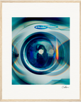 A framed photograph of a front-loading Electrolux washing machine, with colorful reflections and lens flare effects across the glass door. This gallery-quality frame makes an elegant choice for inspirational wall art, signed at the bottom right corner.
