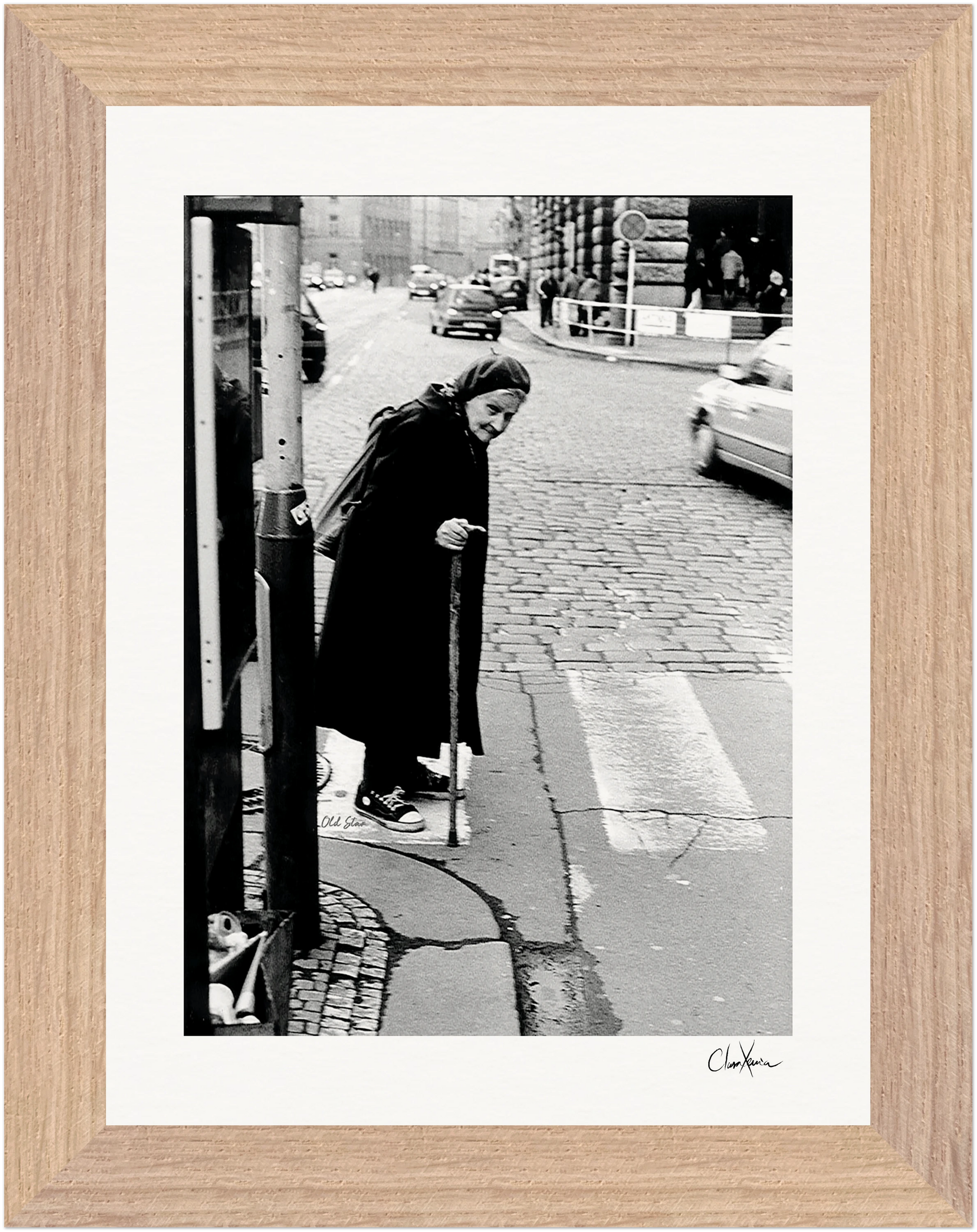 An elderly person in a long coat and headscarf uses a cane while crossing a cobblestone street at a crosswalk. Cars and buildings are visible behind them. This black-and-white fine art print is beautifully presented in a gallery-quality frame.