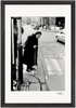 An elderly woman with a cane crosses a cobblestone street in this black and white fine art print. She wears a long coat and headscarf, with cars and buildings in the background—a thoughtful piece for conscious interior design.