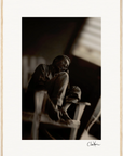 A sepia-toned photograph of an elderly man sitting barefoot on a wooden chair, resting his chin on his knees in a dimly lit room with soft light from a window and framed wall art enhancing the conscious interior design.