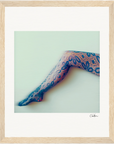 A single leg wearing floral-patterned lace tights is extended against a pale background. This fine art print, with its soft, slightly blurred effect, is framed in light wood—a mindful home decor accent with a small signature in the bottom right corner.