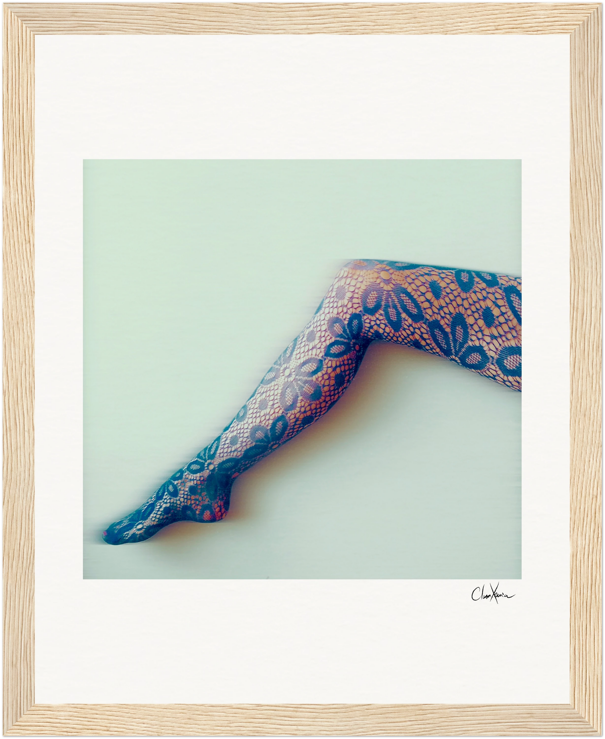A single leg wearing floral-patterned lace tights is extended against a pale background. This fine art print, with its soft, slightly blurred effect, is framed in light wood—a mindful home decor accent with a small signature in the bottom right corner.