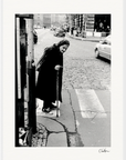 An elderly woman in a headscarf and dark clothing uses a cane to cross a cobblestone street, with cars and buildings in the background—an evocative urban moment, perfect for framed wall art or conscious interior design.