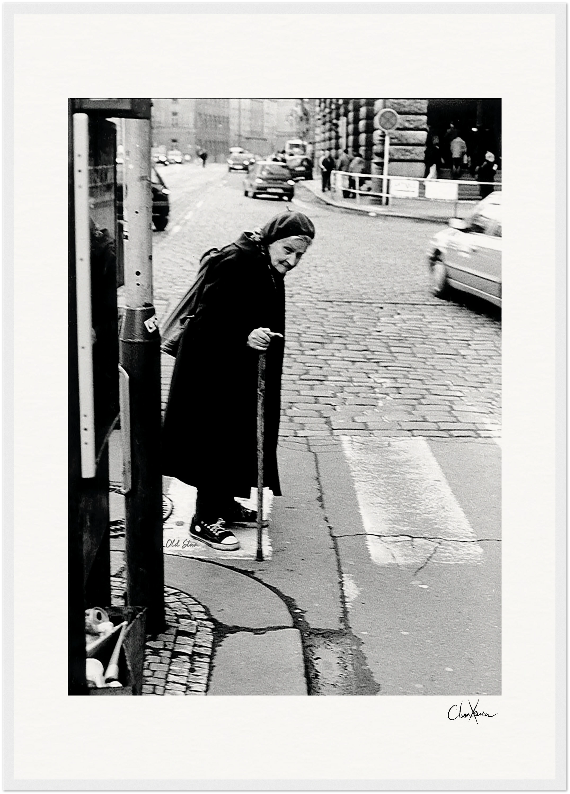 An elderly woman in a headscarf and dark clothing uses a cane to cross a cobblestone street, with cars and buildings in the background—an evocative urban moment, perfect for framed wall art or conscious interior design.