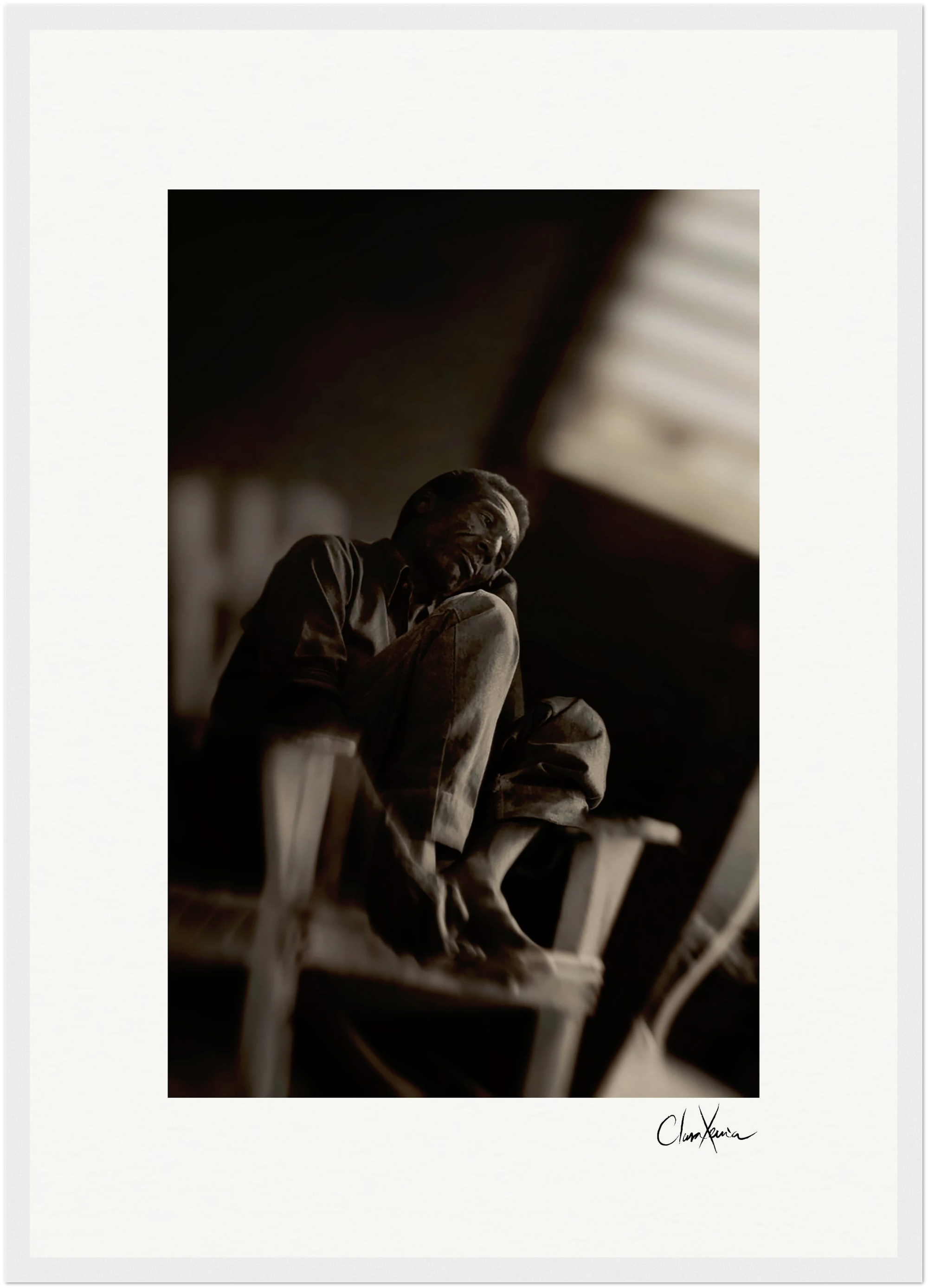 A man sits barefoot on a wooden chair in a dimly lit room, resting his cheek on his hand. This black and white fine art print is signed “Chan Xaru” at the bottom right corner, perfect as framed wall art for sophisticated interiors.