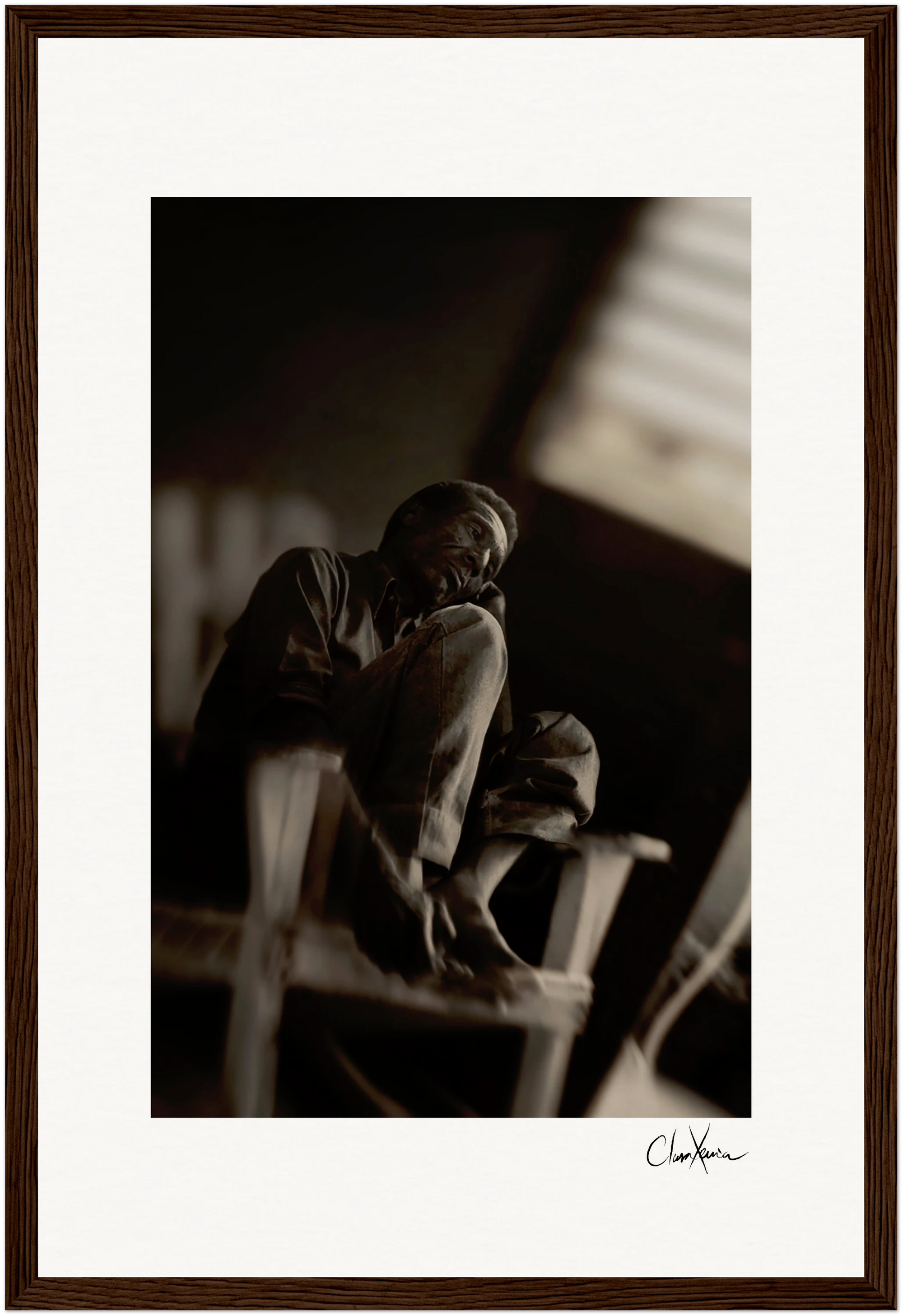 A black-and-white photo of an older man sitting on a wooden chair with one knee up, gazing slightly upward. The softly blurred background enhances the scene, elegantly framed with a gallery-quality frame for conscious interior design.