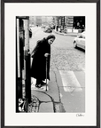 An elderly woman with a headscarf and long coat walks with a cane at a crosswalk on a city street. Cars and buildings appear in the background. This black and white photo is presented as a fine art print in a gallery-quality frame.