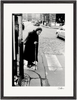 An elderly woman with a headscarf and long coat walks with a cane at a crosswalk on a city street. Cars and buildings appear in the background. This black and white photo is presented as a fine art print in a gallery-quality frame.