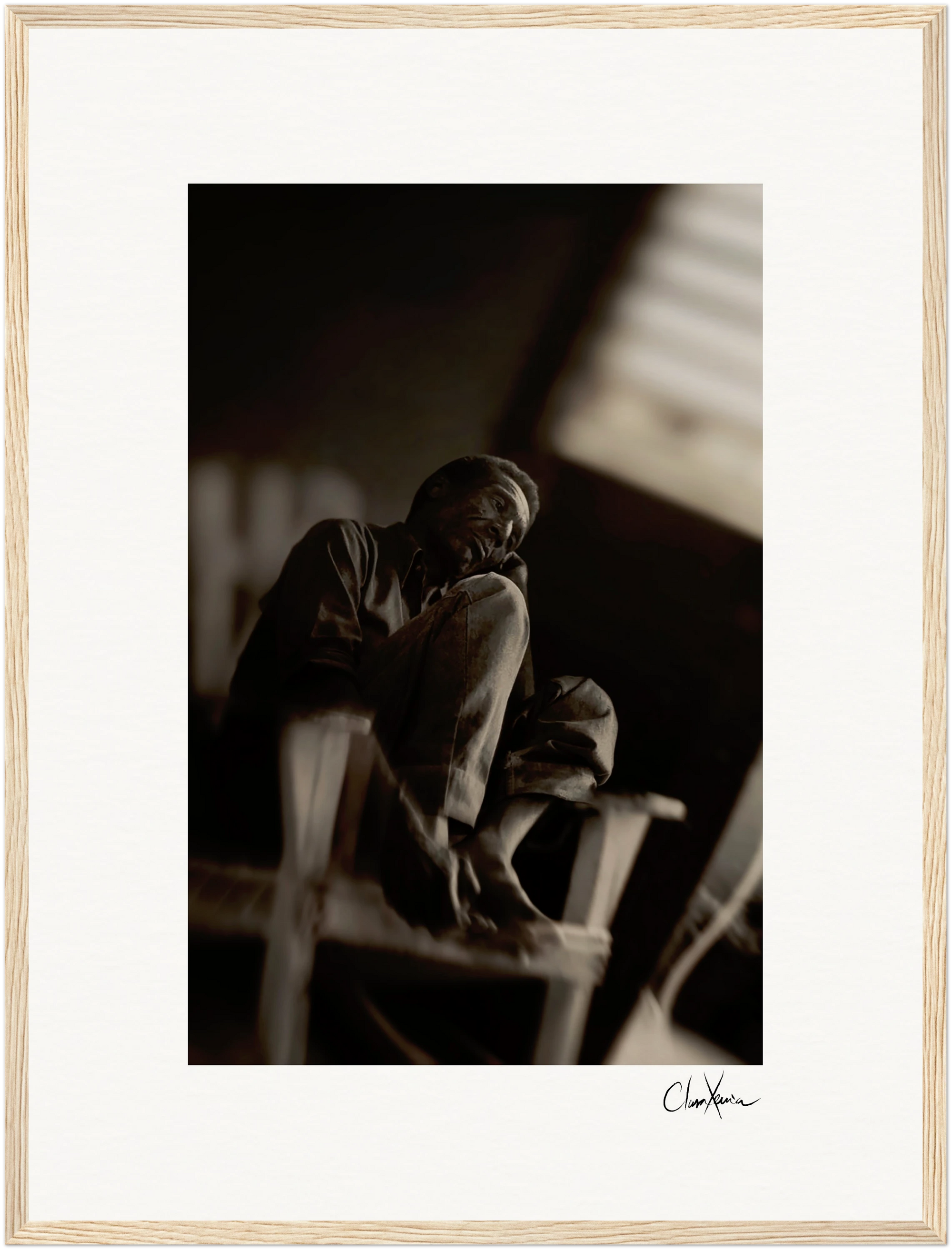 A man sits barefoot on a wooden chair with one knee raised, gazing thoughtfully to the side in a dimly lit room. This fine art print is framed with a white mat and light wood frame, featuring a signature at the bottom—perfect framed wall art.