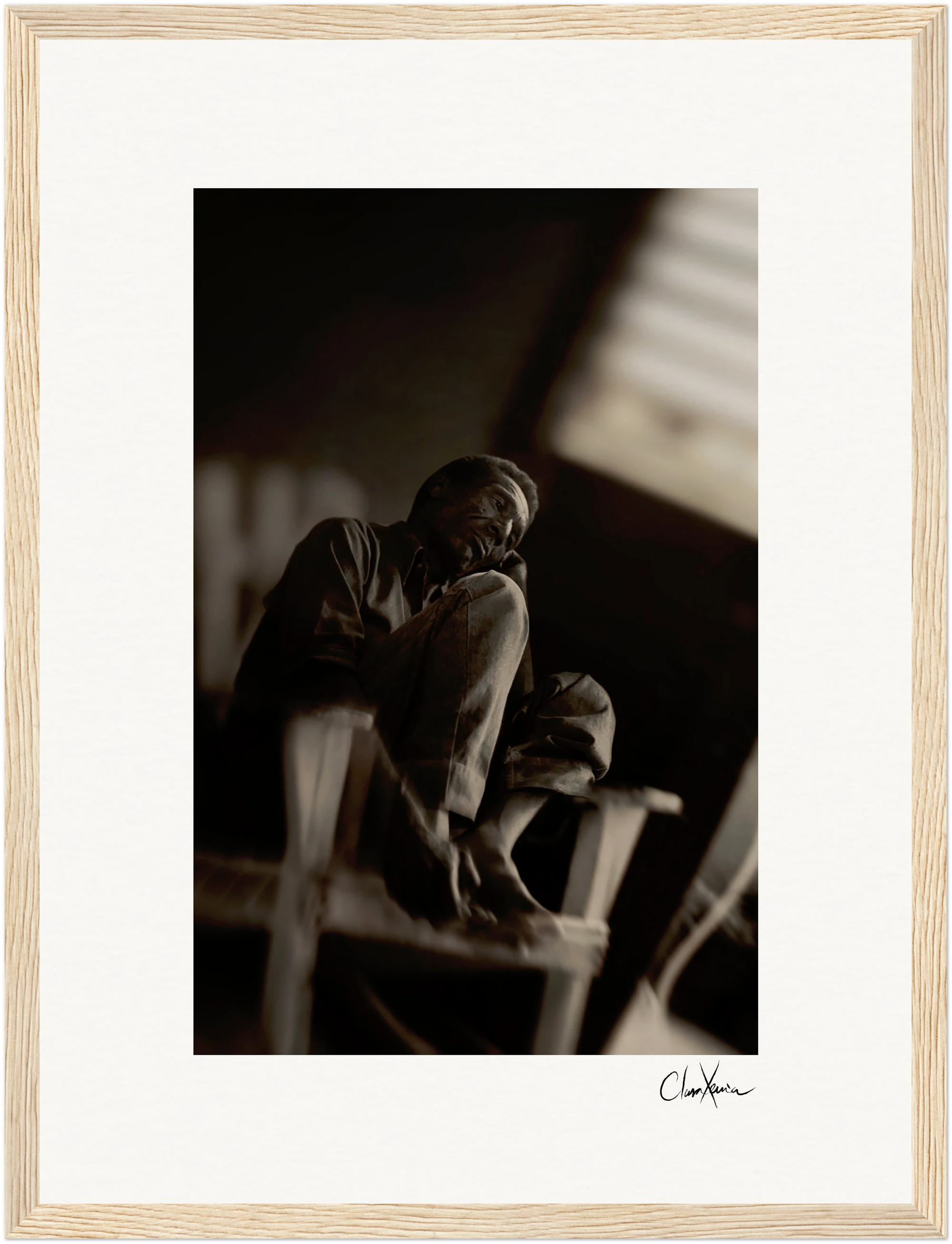 A black-and-white photo shows a person sitting barefoot on a chair with one knee raised, gazing thoughtfully into the distance. The background is blurred, and the image is framed in a gallery-quality frame for conscious interior design.