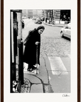 An elderly woman in a headscarf and dark clothing crosses a cobblestone street with a cane. Cars and buildings appear in the background. The black-and-white image is elegantly framed with a gallery-quality frame, perfect for mindful home decor.