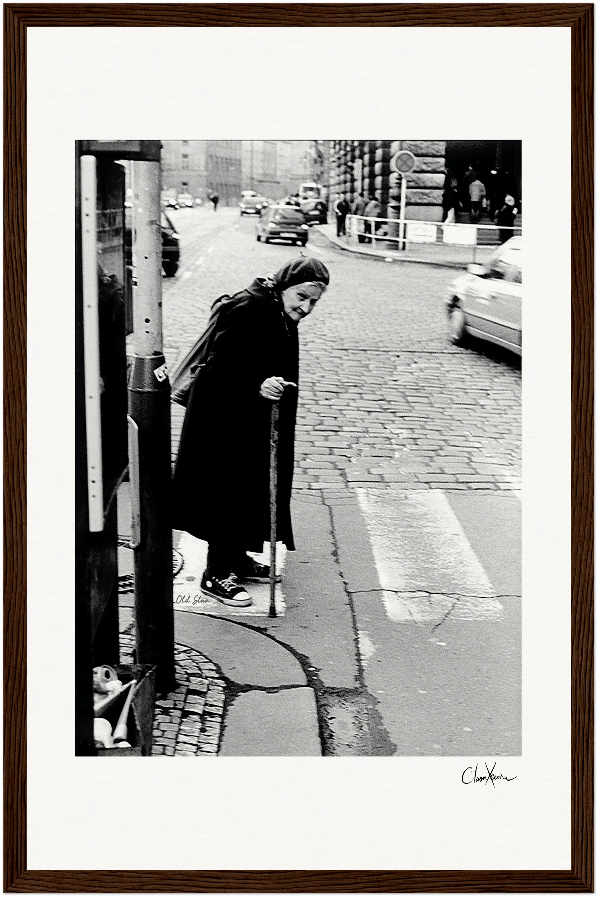 An elderly woman in a headscarf and dark clothing crosses a cobblestone street with a cane. Cars and buildings appear in the background. The black-and-white image is elegantly framed with a gallery-quality frame, perfect for mindful home decor.