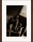 A black and white photo shows an older person sitting on a chair with one knee drawn up, gazing off to the side. Soft, diffused light from a window enhances this fine art print, framed with a dark wooden border—perfect for mindful home decor.